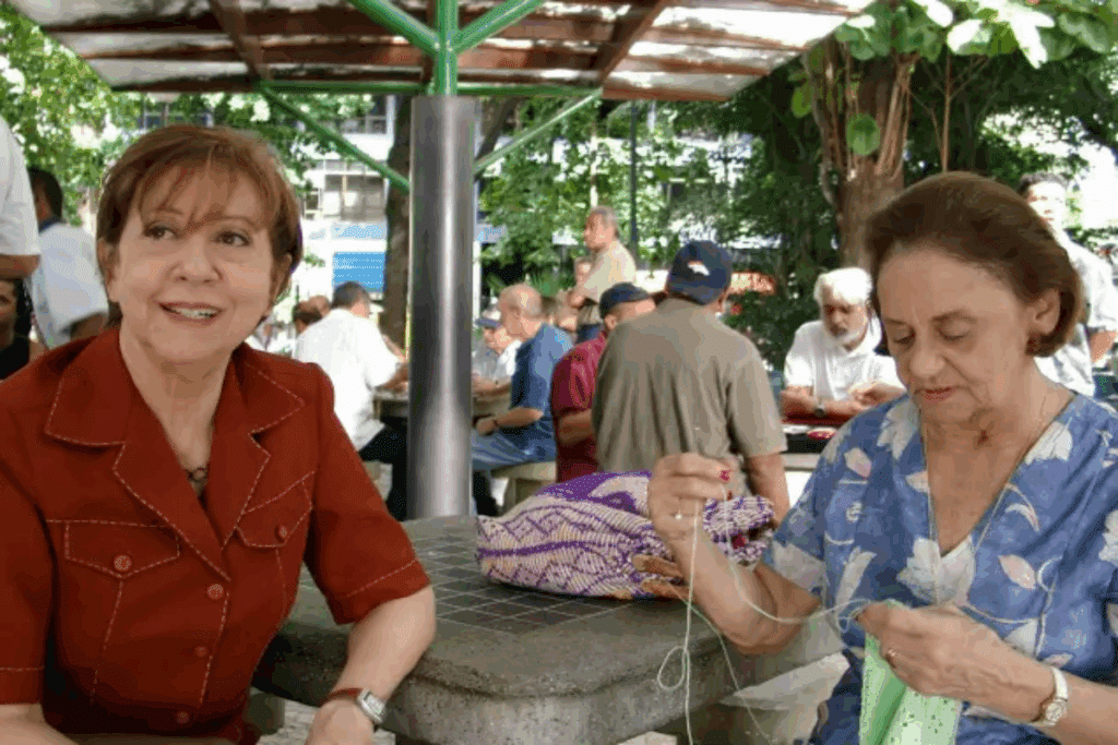 Duas mulheres sentadas em praça com mais pessoas, uma delas tem o cabelo curto e está usando camisa vermelha e a outra tem o cabelo curto, está usando uma camisa azul estampada e está fazendo crochê