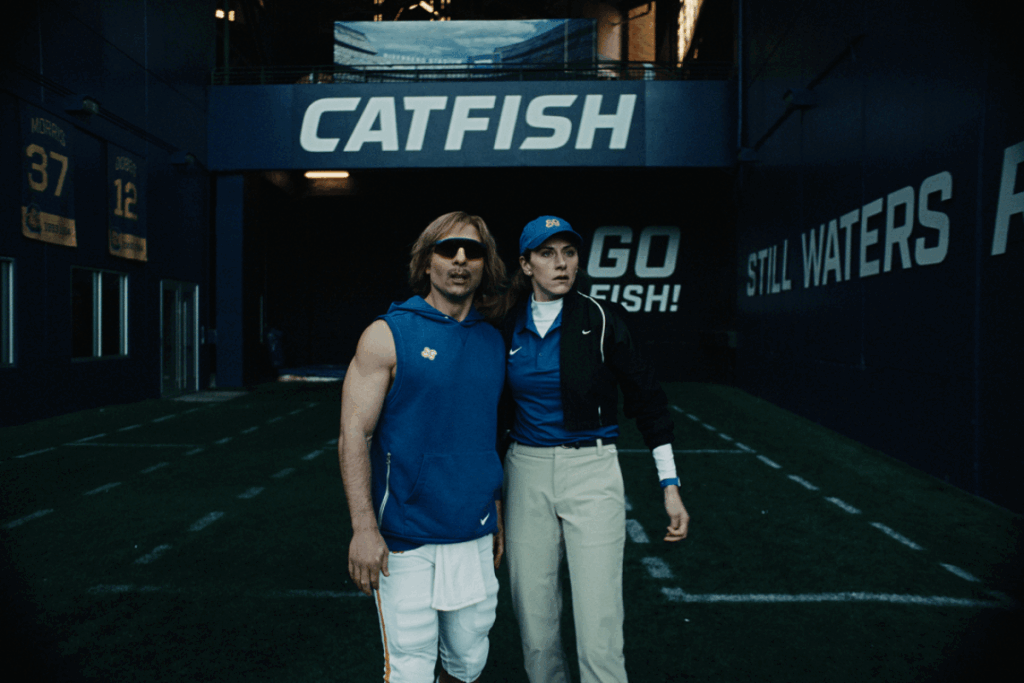 Homem com cabelo loiro, óculos escuro, regata azul e calça branca ao lado de mulher usando boné azul, camiseta azul e branca, calça bege e casaco preto em estádio de futebol americano