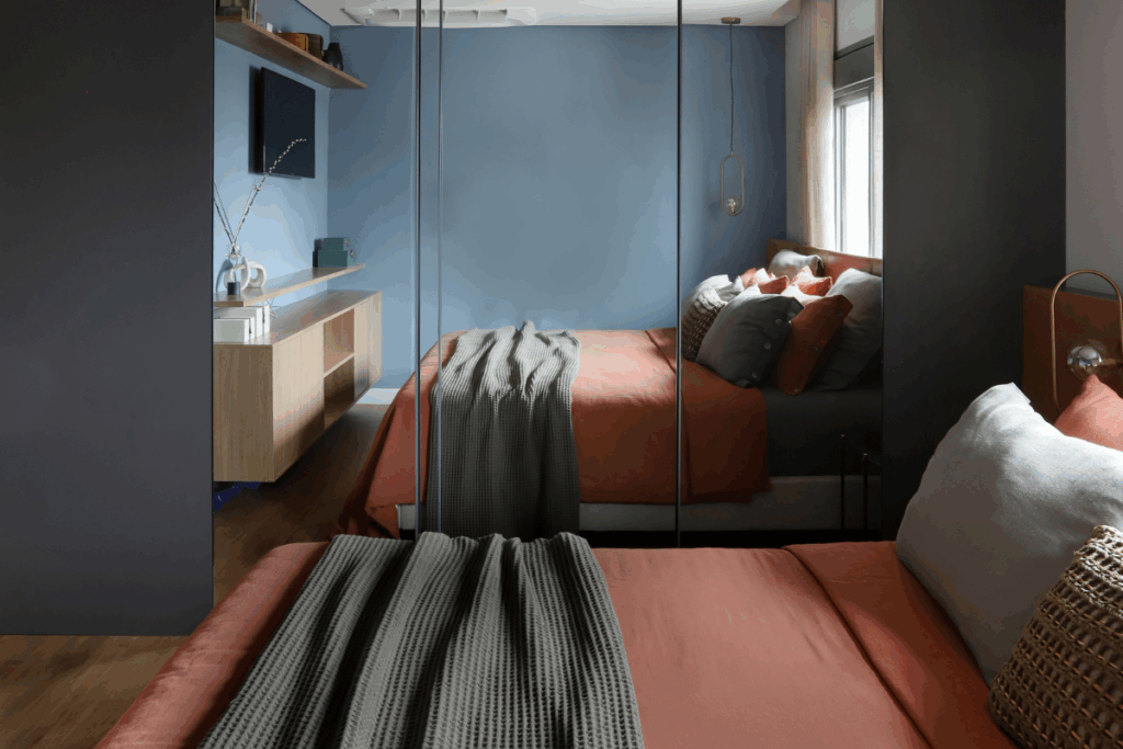 Quarto moderno com paredes pintadas de azul e guarda-roupa espelhado refletindo a cama. A cama possui roupa de cama em tom terracota e uma manta cinza sobreposta. Ao lado, há um móvel suspenso de madeira clara com televisão fixada na parede.