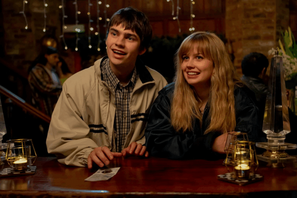 Um jovem casal sentado lado a lado em um balcão de bar iluminado por velas e luzes decorativas. Ambos sorriem enquanto olham para frente.