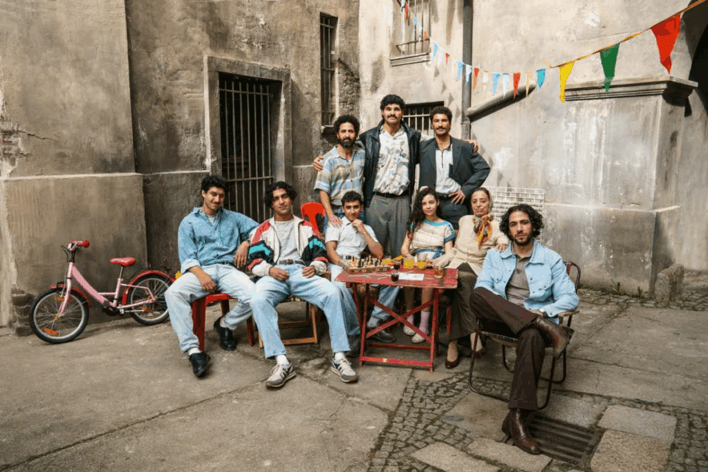 Fotografia horizontal de um grupo de pessoas reunido em um pátio antigo, com paredes desgastadas e bandeirinhas coloridas. Algumas pessoas estão sentadas em cadeiras ao redor de uma mesa, enquanto outras permanecem em pé atrás. Há bebidas e objetos sobre a mesa e uma bicicleta infantil rosa ao lado. A cena transmite um momento de convivência familiar