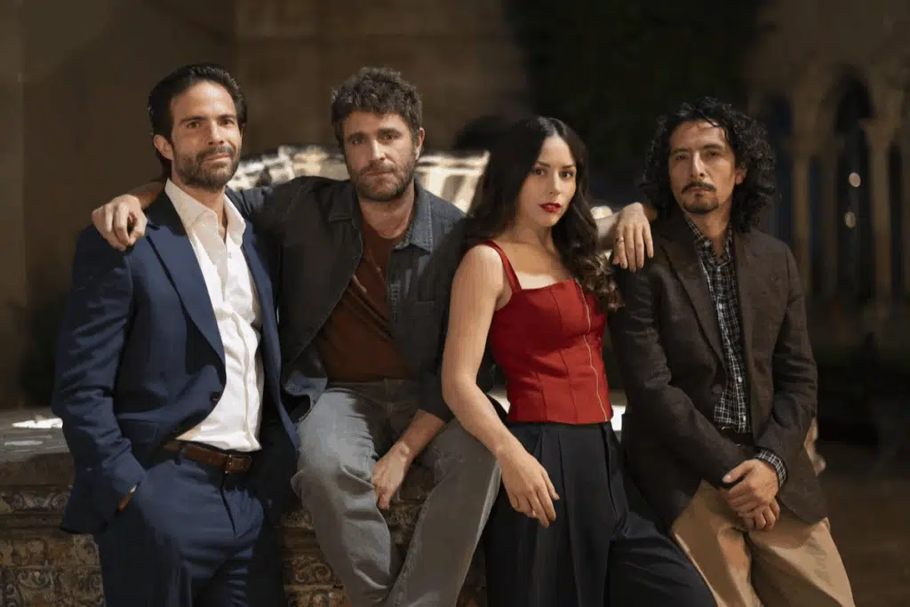Quatro pessoas bem vestidas estão lado a lado ao ar livre, três homens e uma mulher, em pose amigável.
