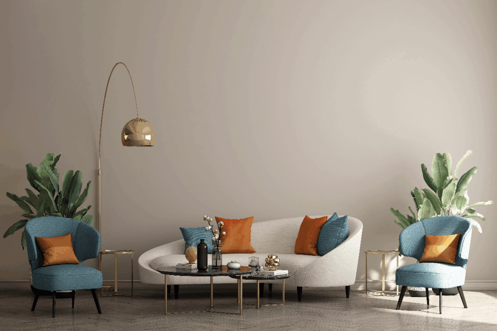 A imagem mostra uma sala de estar elegante, com um sofá branco de formato arredondado, almofadas azuis e alaranjadas e duas poltronas azuis nas laterais. No centro, há mesas de centro com tampo escuro e detalhes dourados. O ambiente também possui plantas verdes, pequenas mesas laterais e uma luminária dourada de chão, sobre um fundo de parede bege.
