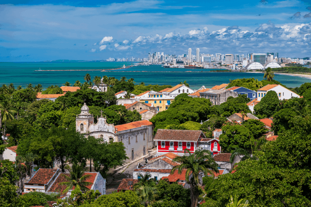 Vista panorâmica do centro histórico de Olinda, em Pernambuco, com igrejas coloniais, casarões coloridos e construções de telhados de barro cercadas por árvores e palmeiras. Ao fundo, destacam-se o mar de águas azul-esverdeadas e os prédios modernos de Recife, sob um céu azul com nuvens brancas.